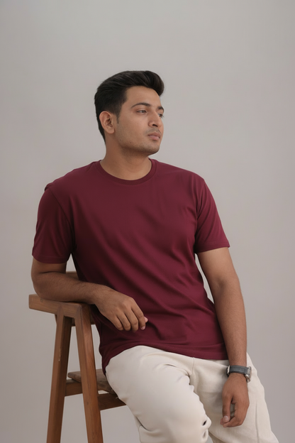 Man wearing a maroon t-shirt and white pants sitting on a wooden stool against a plain background