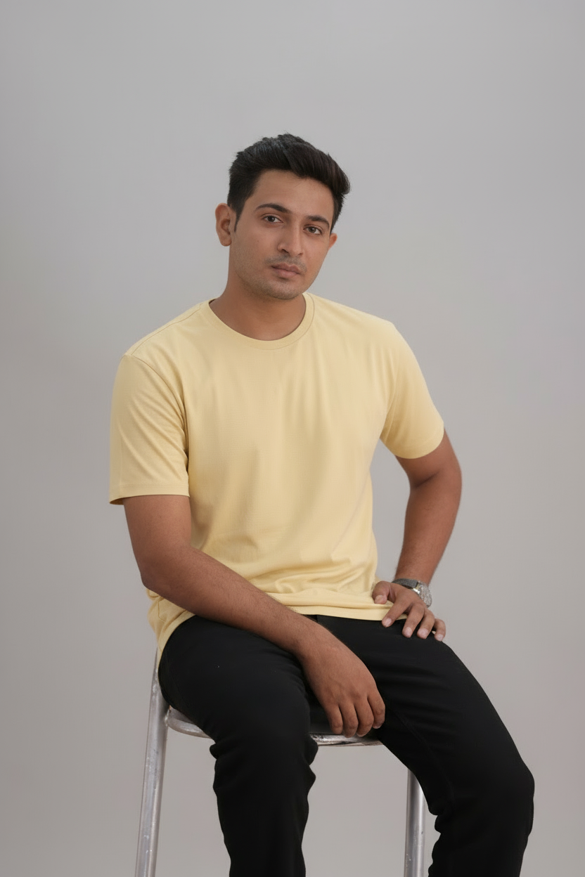Man sitting on a stool wearing a yellow t-shirt and black pants against a gray background